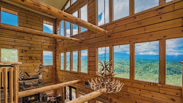 A cabin with a stone fireplace, wooden beams, and large windows offering a view of the forest. - PinnacleView: POOL, Breathtaking Views, Movie Room, Games, Hot Tub, Wifi (Sevierville)