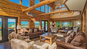 A spacious wooden interior with multiple seating areas, large windows offering scenic views, and a fireplace. - PinnacleView: POOL, Breathtaking Views, Movie Room, Games, Hot Tub, Wifi (Sevierville)