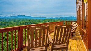 Um deck de madeira com cadeiras, com vista para uma cadeia montanhosa arborizada.