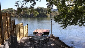 Een houten terras met tafel en stoelen met uitzicht op een rustig meer, een steiger en een lantaarnpaal.
