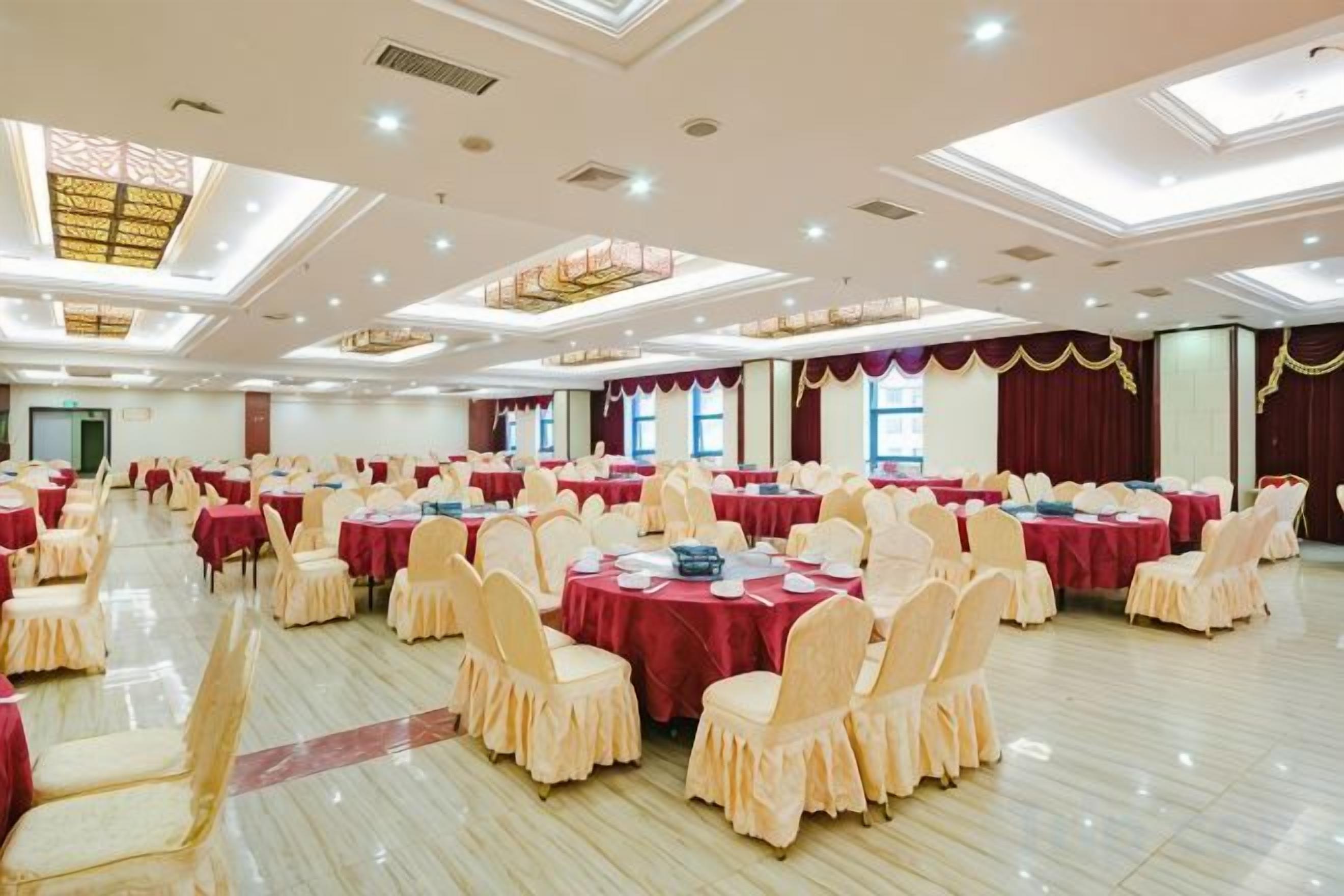 A banquet hall with multiple round tables, each covered with a red tablecloth, surrounded by chairs with a light-colored cover. The room has a high ceiling with recessed lighting and a patterned ceiling design.