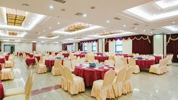 A banquet hall with multiple round tables, each covered with a red tablecloth, surrounded by chairs with a light-colored cover. The room has a high ceiling with recessed lighting and a patterned ceiling design.