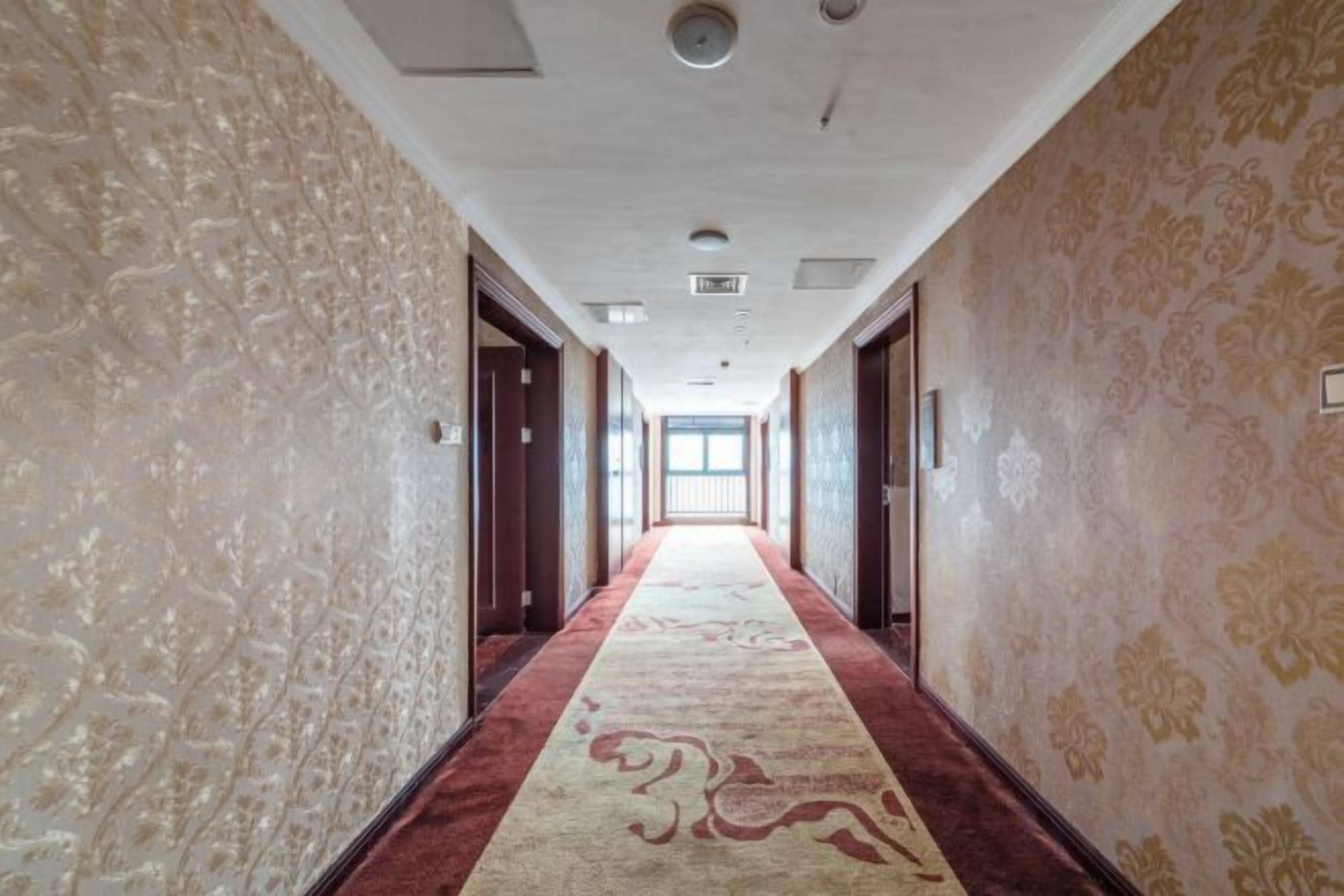 Un couloir d'hôtel aux murs ornés de motifs, avec un tapis rouge et des portes donnant accès aux chambres.