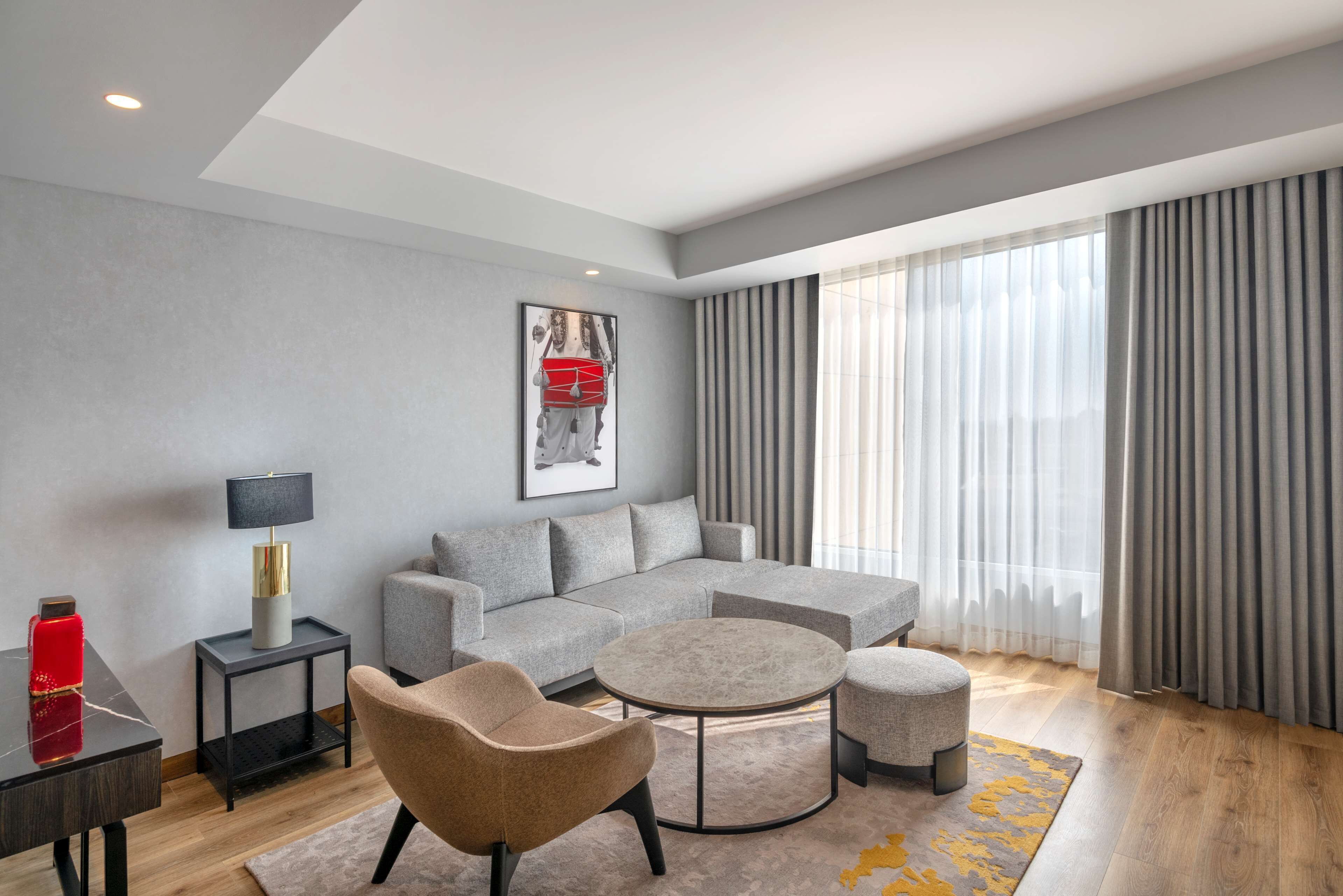 A modern living room with a grey sofa, a round coffee table, and a beige armchair. There is a small side table with a lamp and a red decorative item. A framed picture hangs on the wall, and the room has large windows with grey curtains.