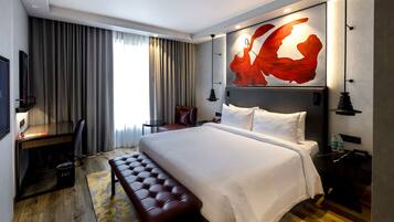 Una habitación de hotel moderna con una cama grande, un escritorio, un banco de cuero, un televisor y un cuadro abstracto de gran tamaño en la pared.
