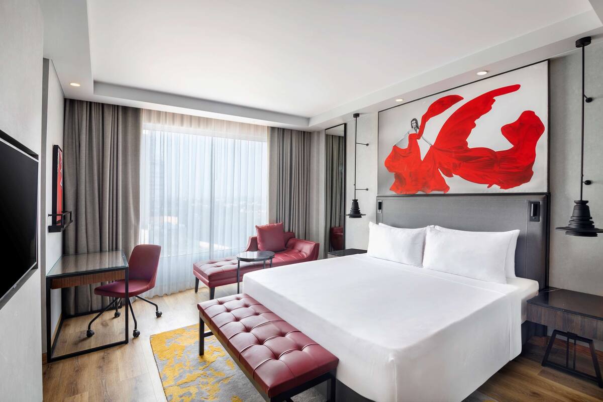 A hotel room with a large bed, a desk, a chair, a bench, and a large window with curtains.