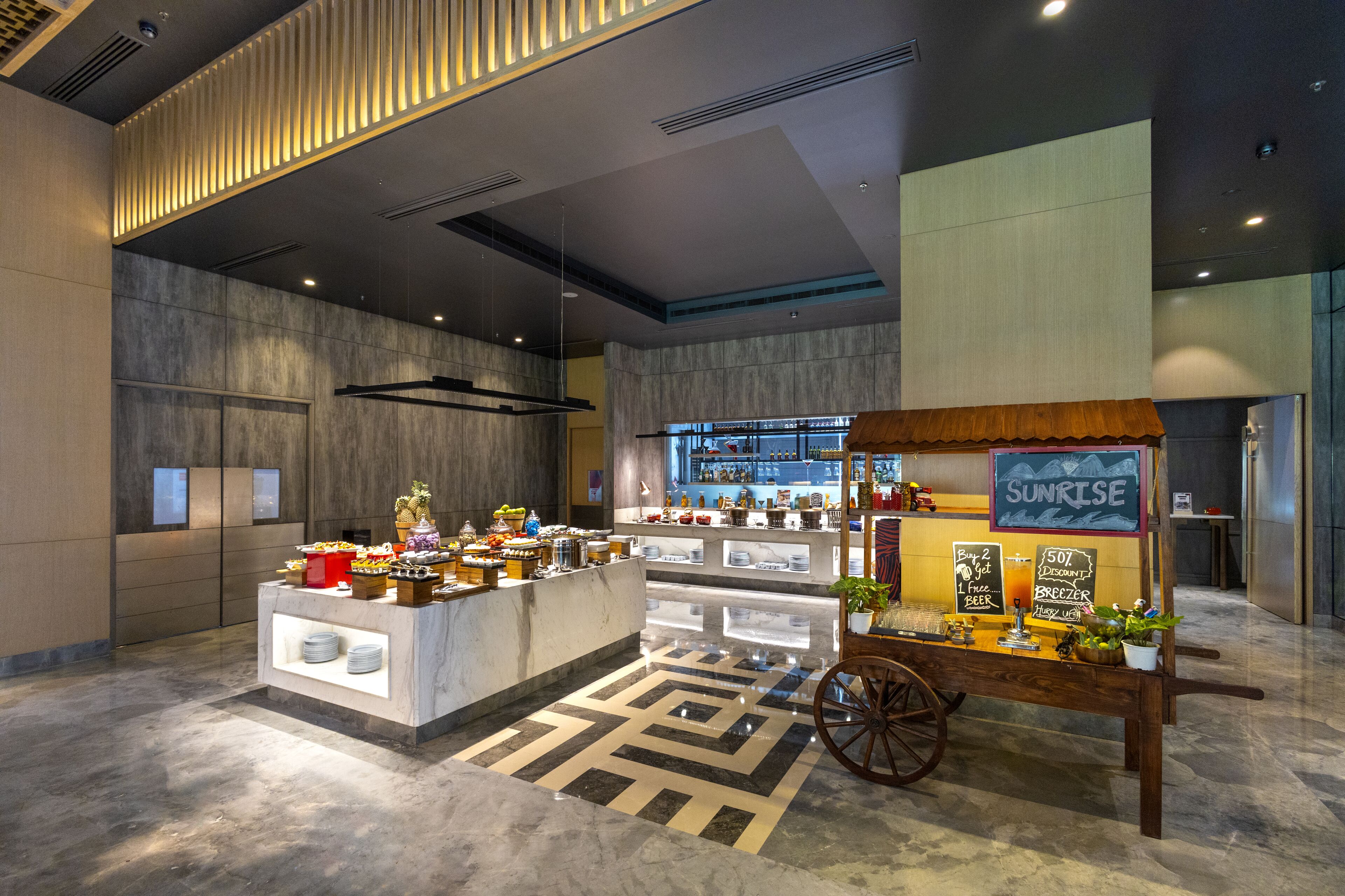 A modern hotel lobby with a breakfast bar, a cart labeled "SUNRISE," and a bar area in the background.