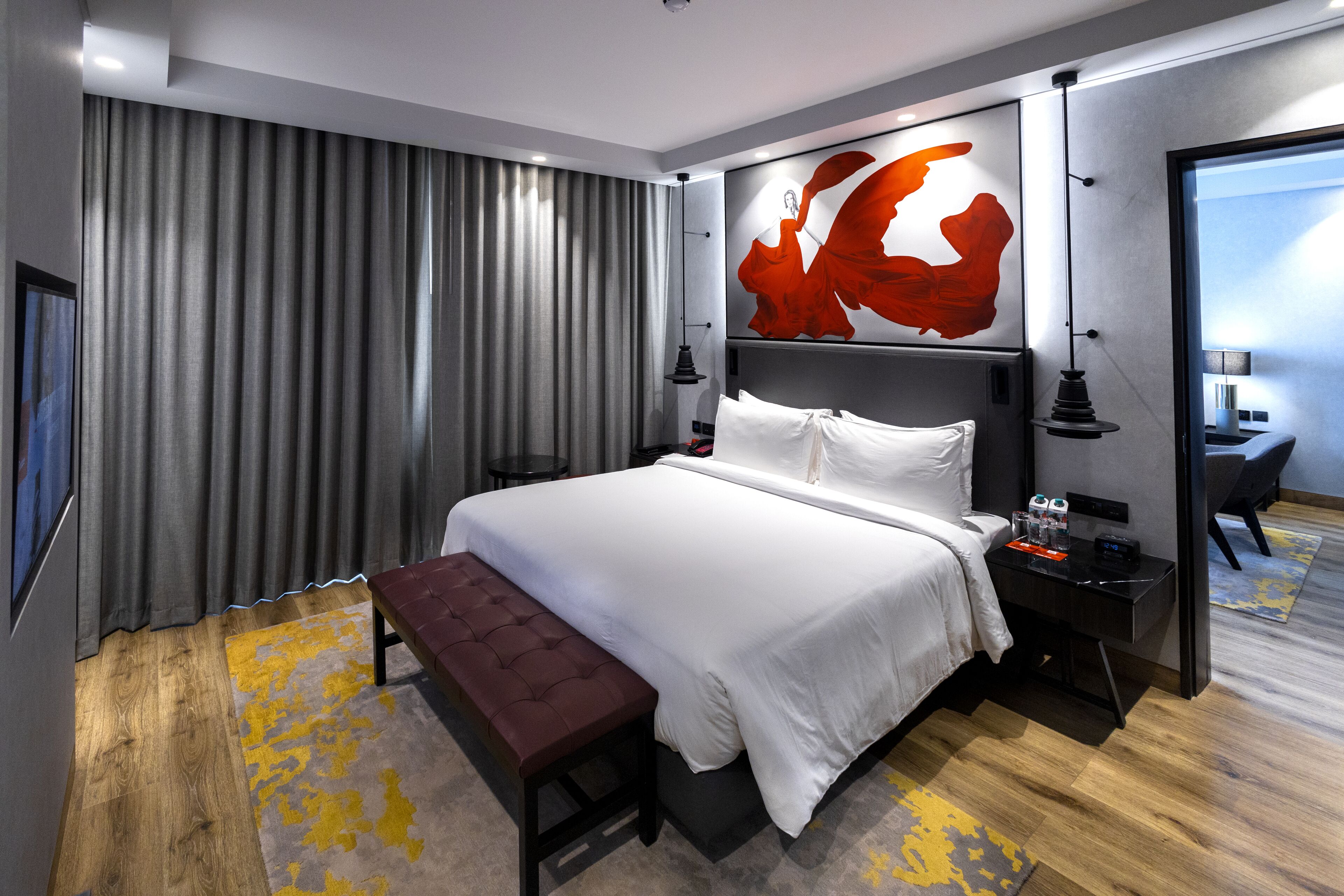 A modern hotel room with a large bed, a bench, a desk, and a painting on the wall.