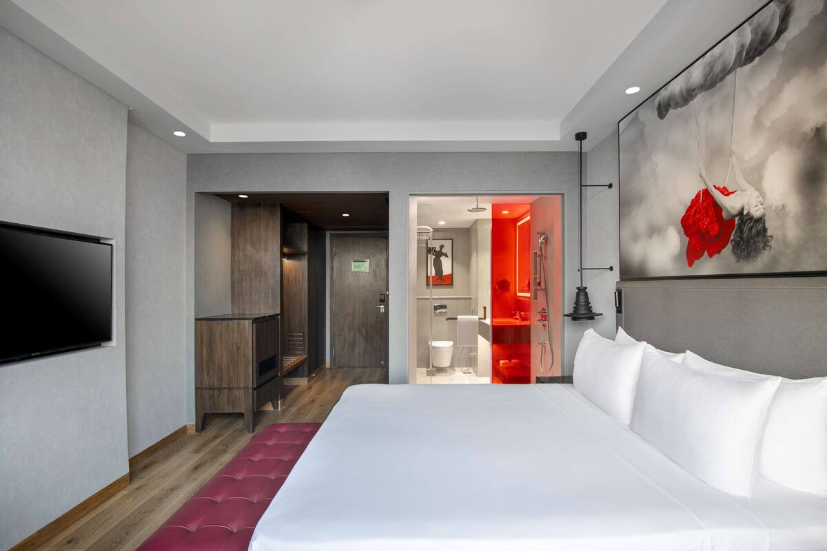 A modern hotel room with a large bed, a flat-screen TV, a wooden bench, and a red accent wall.