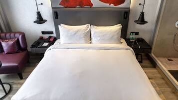 Habitación de hotel con una cama grande, un banco, un cuadro abstracto rojo en la pared, una silla roja y una mesita con un teléfono y una botella.