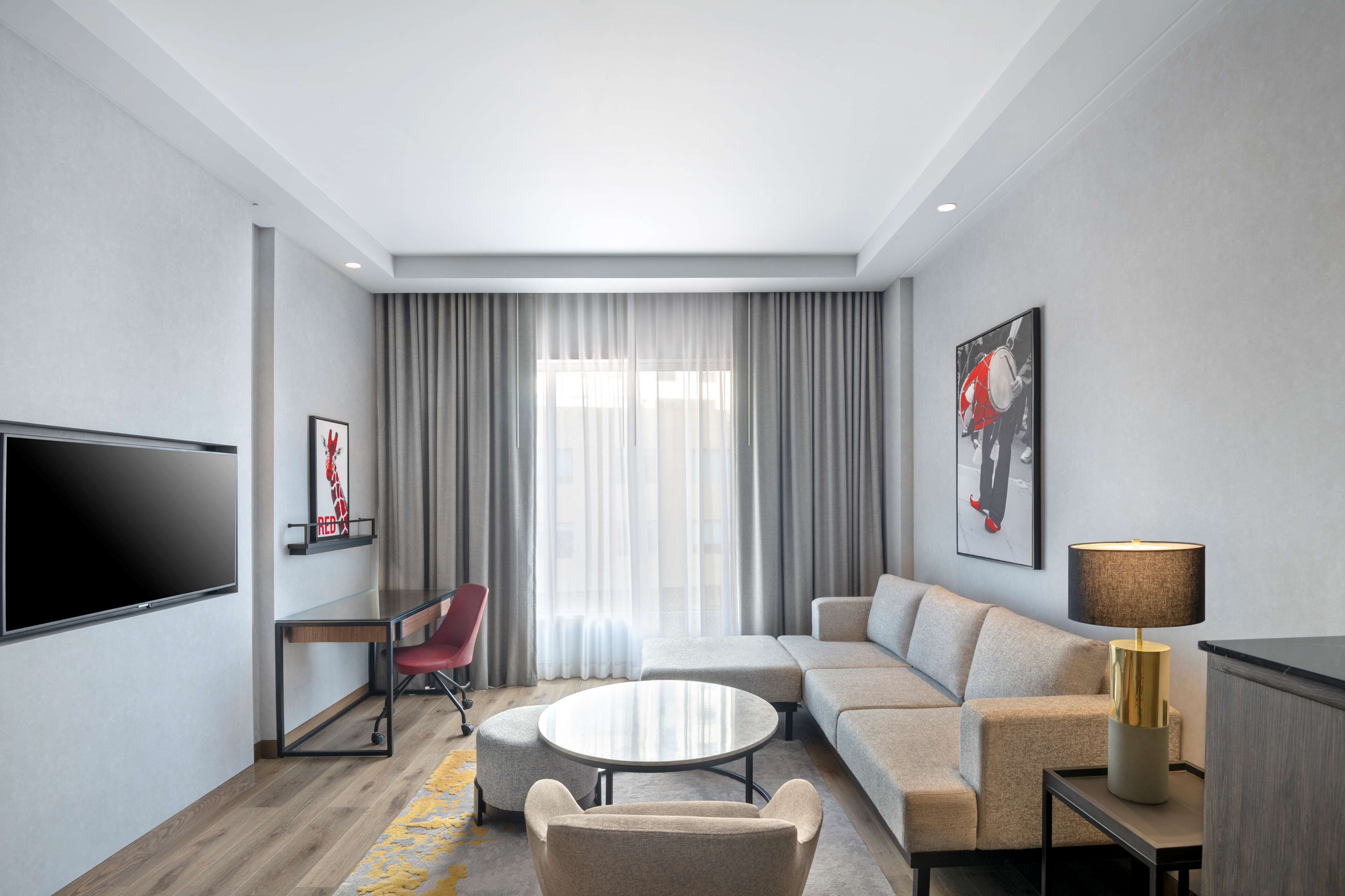 A modern hotel room with a flat-screen TV, a sofa, a round coffee table, a red chair, and a lamp.
