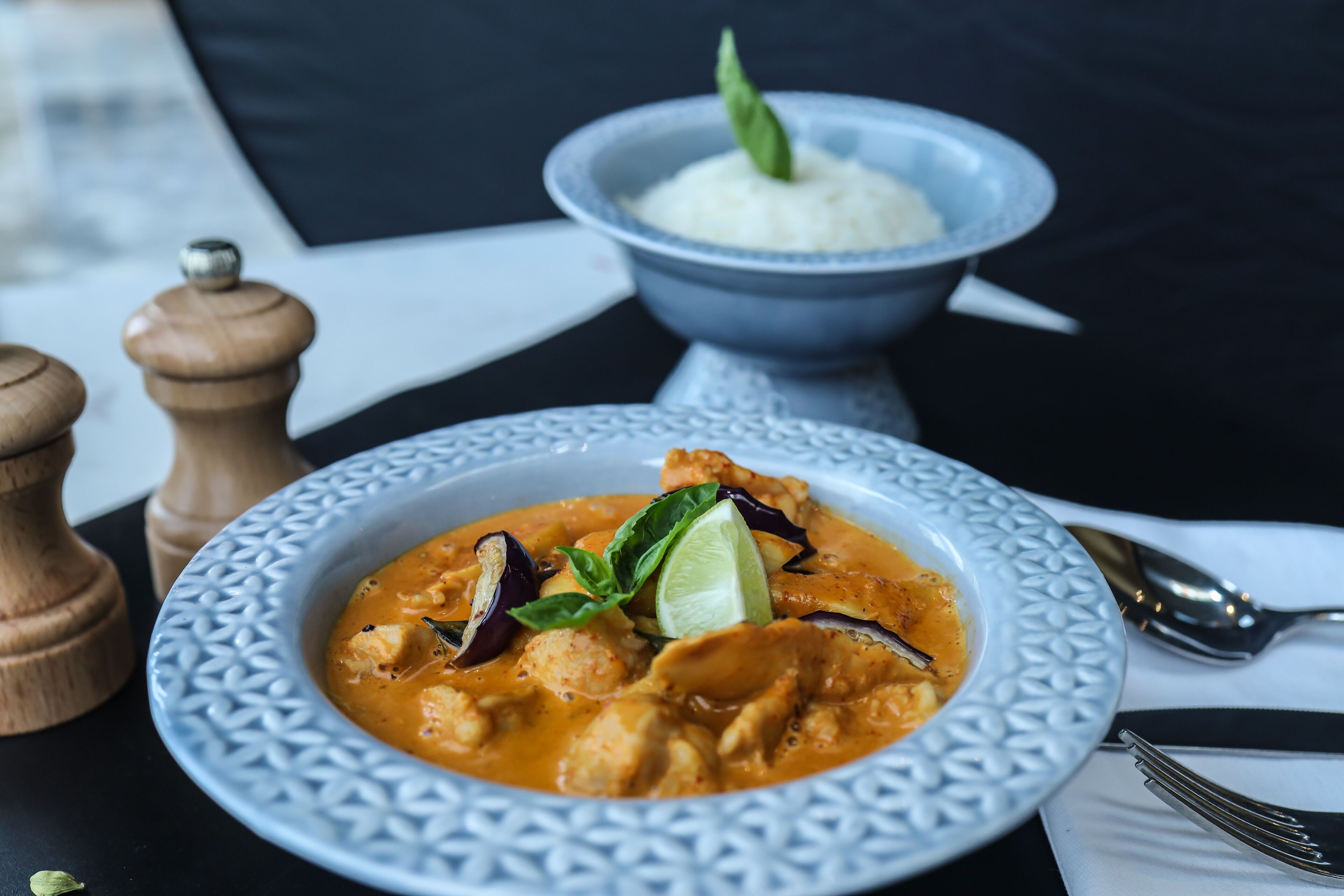 Un plato de curry con lima y albahaca, un cuenco de arroz y un molinillo de pimienta sobre una mesa.