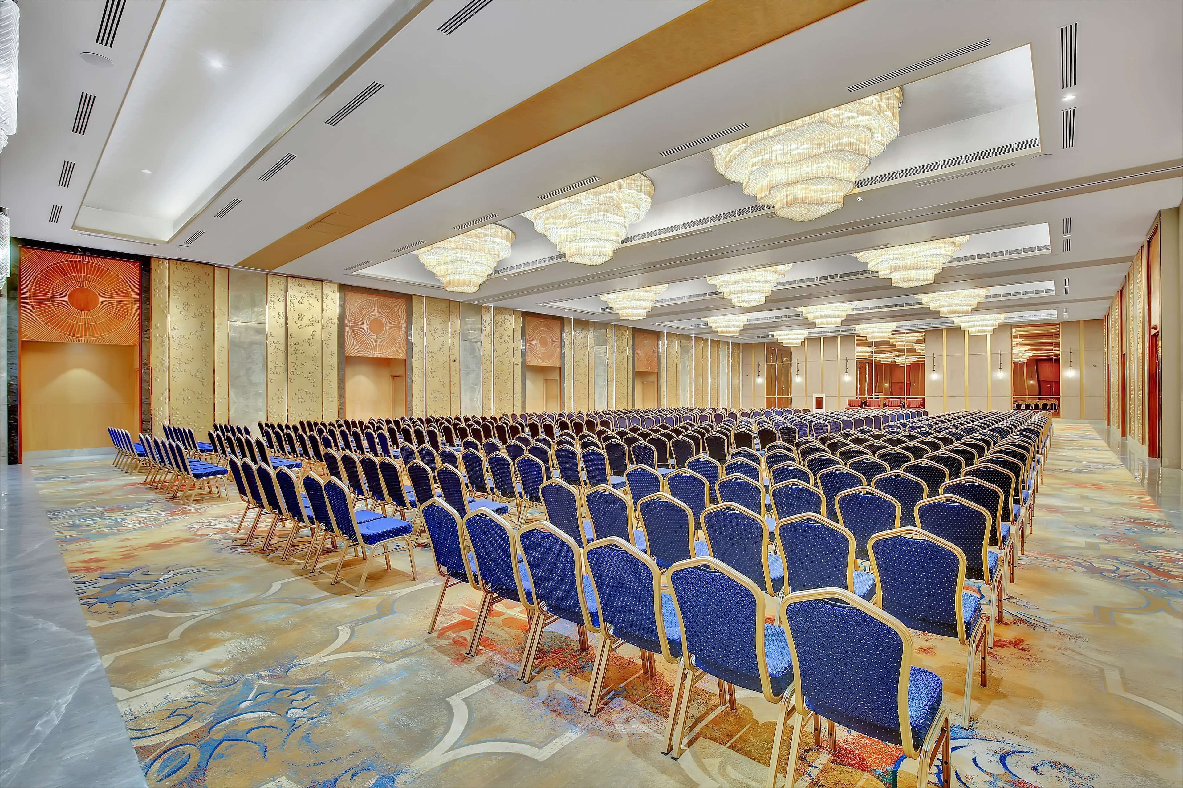 Un amplio salón de conferencias con filas de sillas azules, una alfombra estampada y modernos candelabros.