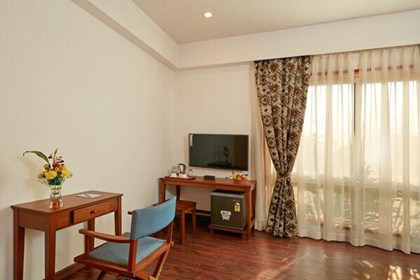 A room with a wooden desk, a television, a small refrigerator, a blue chair, and a window with patterned curtains.