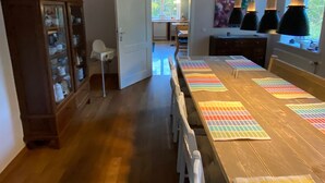 Un espace repas avec une longue table en bois, des chaises blanches et des lampes suspendues. Un buffet en bois avec des portes en verre est visible sur la gauche.