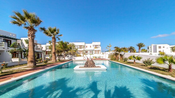 A swimming pool with a rock centerpiece, surrounded by palm trees and white buildings.