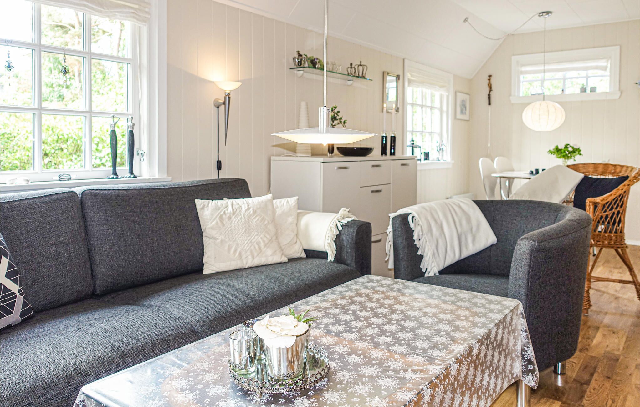 A modern living room with a grey sofa, a coffee table with a floral tablecloth, and a dining area with wicker chairs.