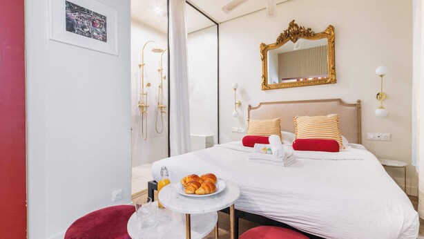 A hotel room with a bed, a bedside table with a plate of croissants, a red chair, a small round table with a glass, and a shower area with a golden showerhead. - Charmant Studio Paris 15e Proche Tour Eiffel (Paris)