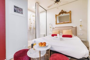 Camera d'albergo con un letto, un comodino con un piatto di croissant, una sedia rossa, un tavolino con un bicchiere e un angolo doccia con soffione dorato.
