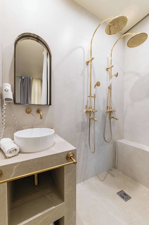 A modern bathroom with a round white sink, gold fixtures, a large mirror, and a shower with gold fixtures.