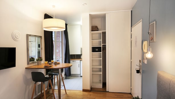Una habitación de hotel compacta con cama, escritorio con sillas, mesa de comedor, armario y pequeña cocina.