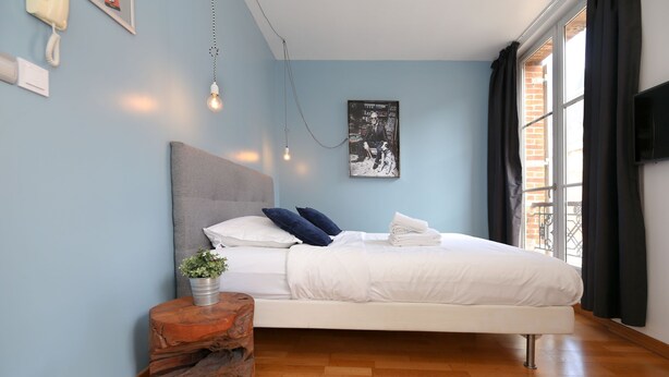Un dormitorio con cama, una mesita de noche con una planta, una lámpara de pared y una ventana con cortinas.
