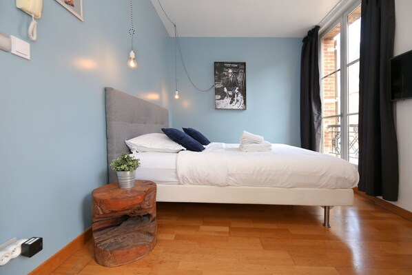 A bedroom with a bed, a bedside table with a plant, a wall-mounted light, and a window with curtains.