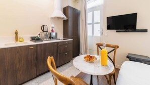 Eine kompakte Küche mit Mikrowelle, Wasserkocher und Kaffeemaschine, einem Esstisch mit Stühlen, einem an der Wand befestigten Fernseher und einem Fenster mit Vorhängen.