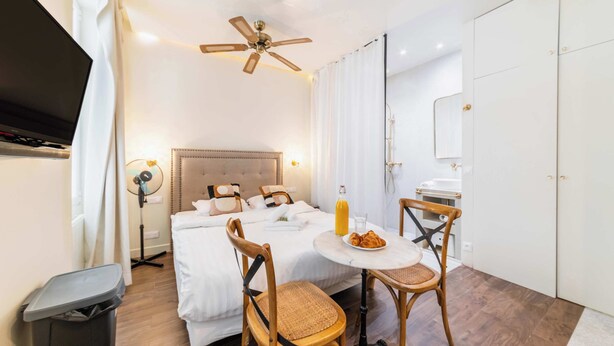 Ein Hotelzimmer mit Bett, Deckenventilator, Tisch mit Flasche und Croissants, Sessel und Fernseher.