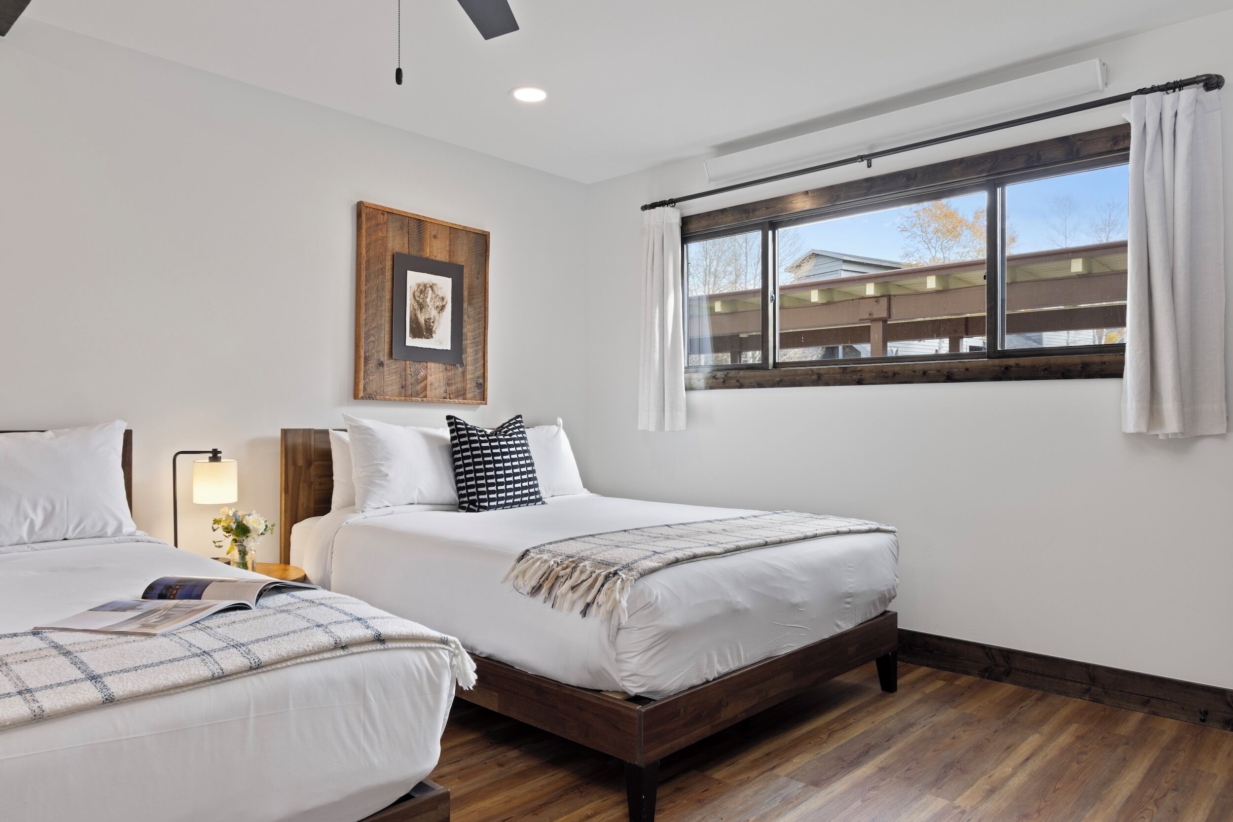 A hotel room with two beds, a large window with curtains, a wooden floor, and a framed picture on the wall.