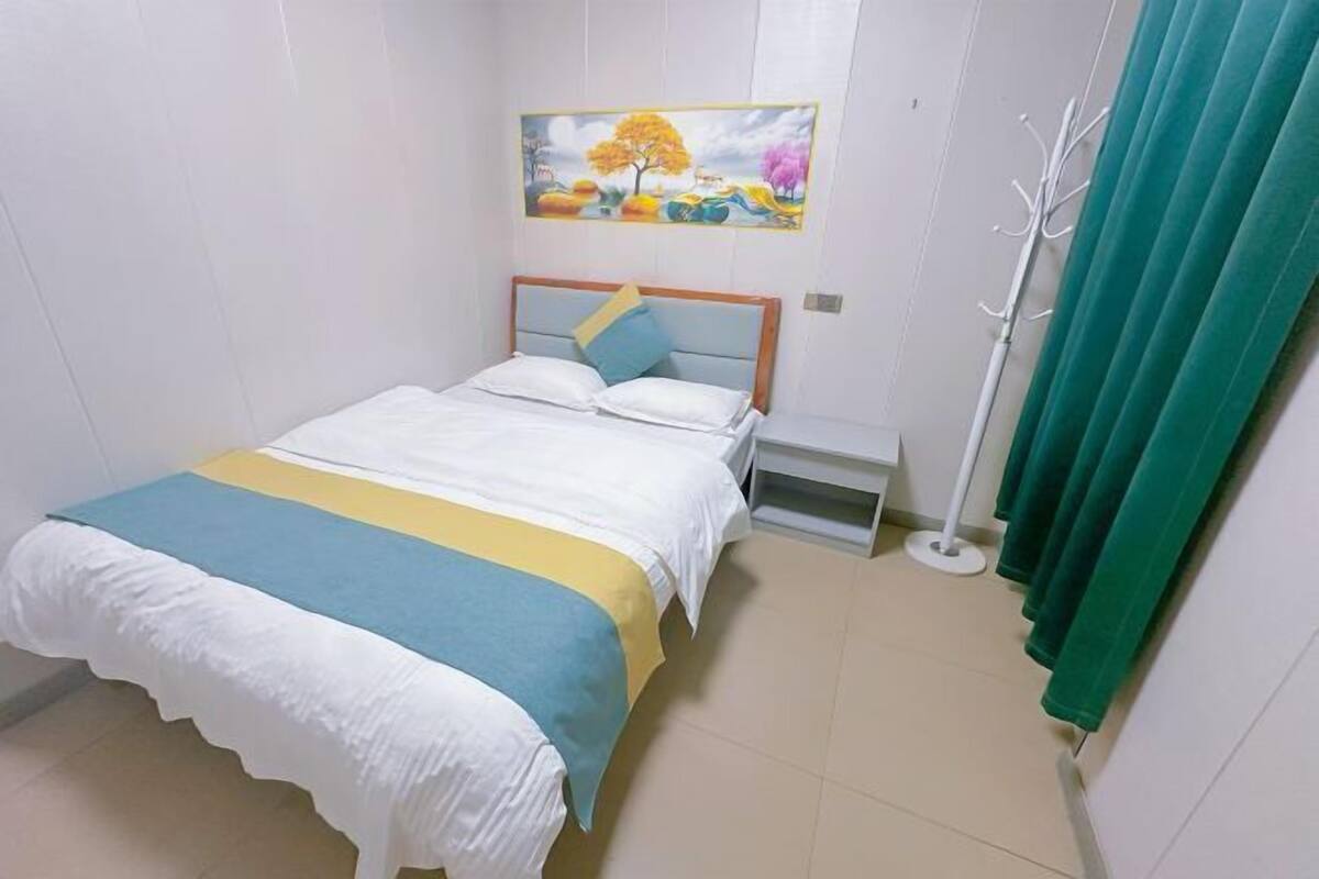 Une chambre à coucher avec un lit, une table de chevet, un tableau au mur, un porte-manteau et un rideau.