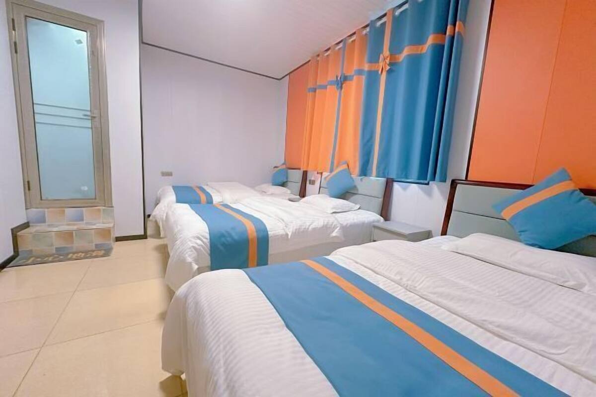 Une chambre d’hôtel avec deux lits, chacun agrémenté de draps blancs et de couvre-lits rayés bleus et oranges. Une grande fenêtre, ornée de rideaux, et une porte donnant sur un balcon complètent la pièce.