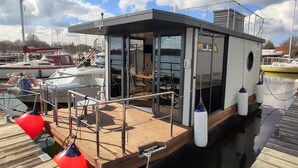 Eine schwimmende Plattform mit überdectem Deck, einem kleinen, abgeschlossenen Bereich mit Tisch und Stühlen sowie Blick auf den Hafen mit anderen Booten.