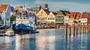Eine Uferlandschaft mit einem blau-weißen Boot, das an einem Steg liegt, umgeben von farbenfrohen Gebäuden mit traditioneller Architektur.