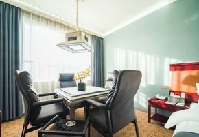 A hotel room with a dining table, two chairs, a small side table with a lamp, and a bed with a red headboard.