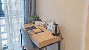 Et hotelværelse med skrivebord, stol, en potteplante, elkedel og et vindue med gardiner.