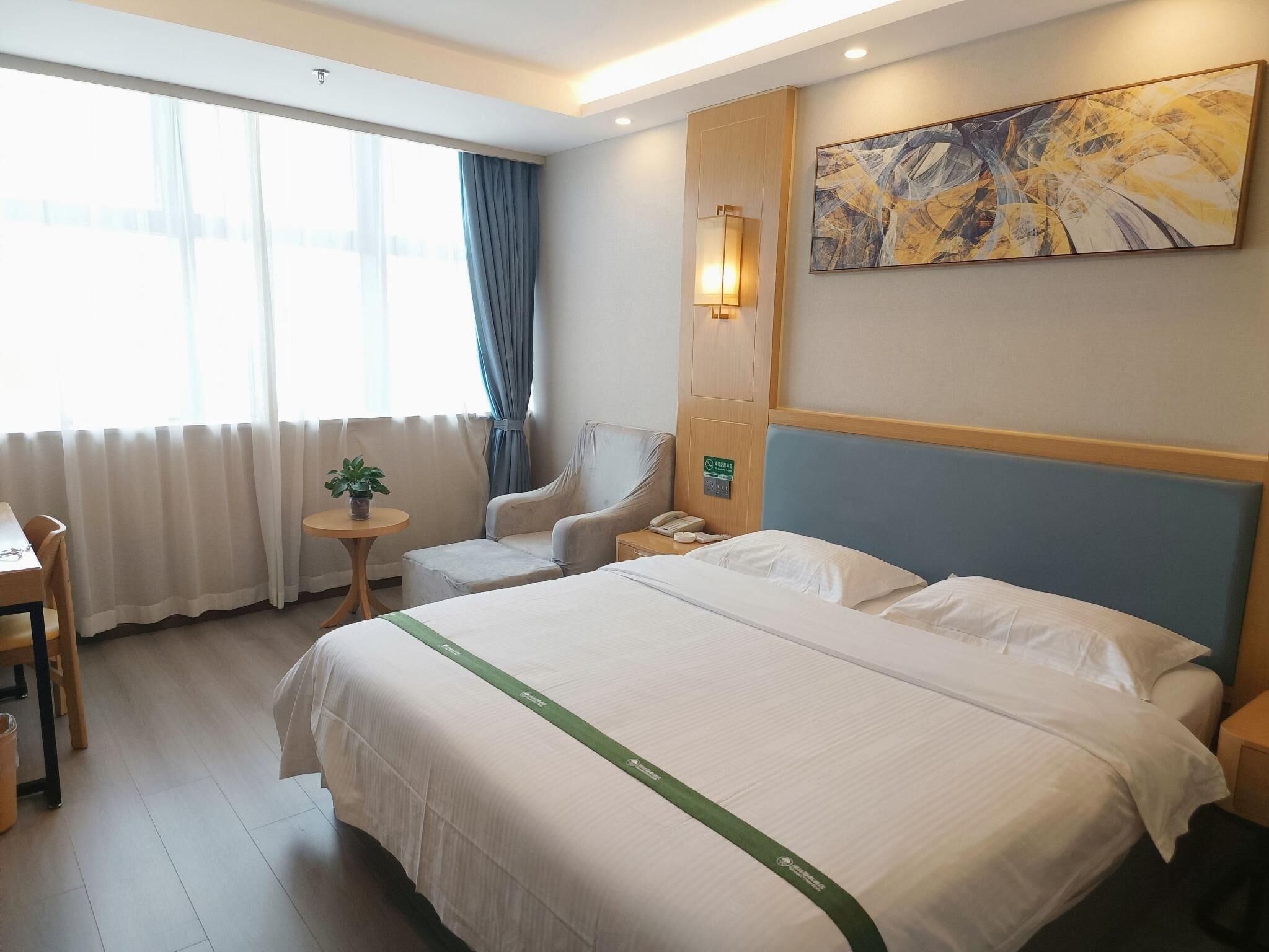 A hotel room with a large bed, a chair, a small table with a plant, a painting on the wall, and a window with curtains.