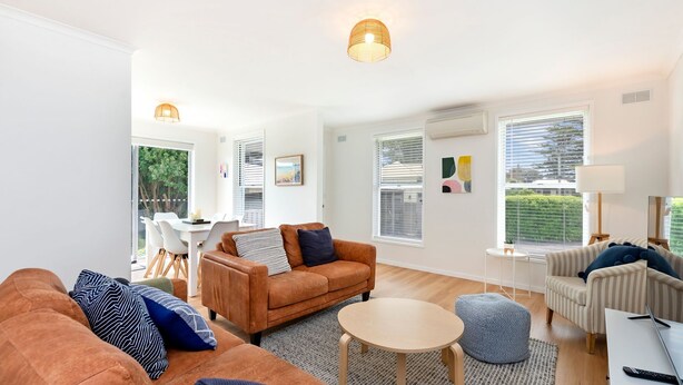 A modern living room with a brown sofa, a round coffee table, and a striped armchair. There are two windows with blinds, a dining area with a white table and chairs, and a small side table with a lamp. - MaryMae - Town Central (Port Fairy)