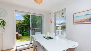 A modern dining area with a white table, chairs, and a view of a garden through glass doors. - MaryMae - Town Central (Port Fairy)