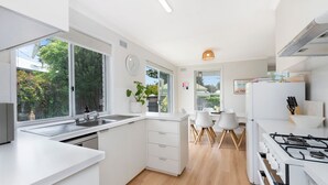 A modern kitchen with white cabinetry, a gas stove, a sink, and a dining area with white chairs. - MaryMae - Town Central (Port Fairy)