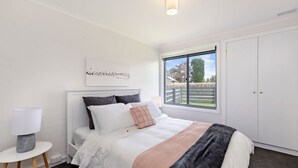A bedroom with a bed, a bedside table, a lamp, a window with a view of the outside, and a wardrobe. - MaryMae - Town Central (Port Fairy)