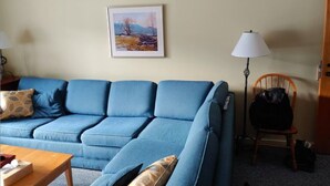 Ein Wohnzimmer mit einem blauen Sofa, einem Holztisch, einem gerahmten Gemälde an der Wand, einer Stehlampe und einem Holzstuhl.