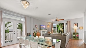 A modern living room with a glass dining table, white chairs, a grey sofa, and a ceiling fan. - Heated Pool, Tiki Lounge, Steps to Beach, Games (St. Augustine)