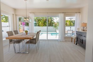 A dining area with a table and chairs, a view of a swimming pool, and a kitchen area with a cabinet and a lamp.