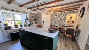A modern kitchen with a central island, wooden beams, pendant lights, and a dining area with a patterned wallpaper backdrop. - No 4 old Post Office Row, Isle of Skye (Skeabost)
