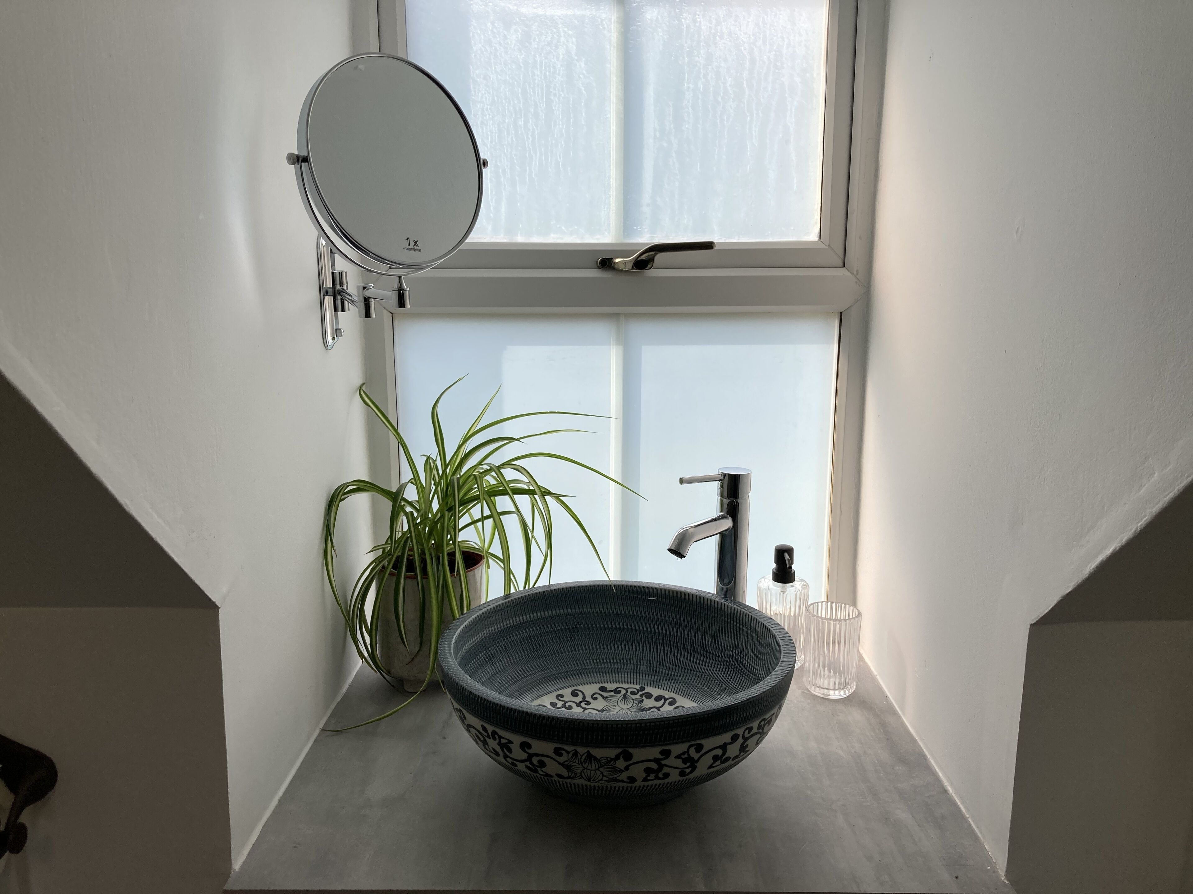 A bathroom with a round mirror, a modern faucet, a decorative bowl sink, a potted plant, and a window with frosted glass.