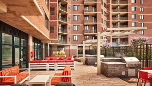 A courtyard with outdoor seating, a barbecue area, and a view of a brick building with balconies. - Sojourn | Courtland Towers | 2 BR (Arlington)