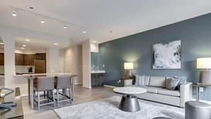 A modern living room with a sofa, coffee table, and chairs. There is a kitchen area with cabinets and a dining table. - Sojourn | Courtland Towers | 2 BR (Arlington)