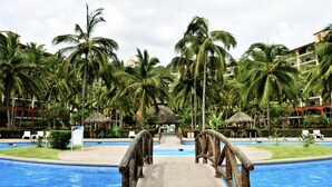A swimming pool with a thatched-roof structure in the center, surrounded by palm trees and a hotel building in the background.
