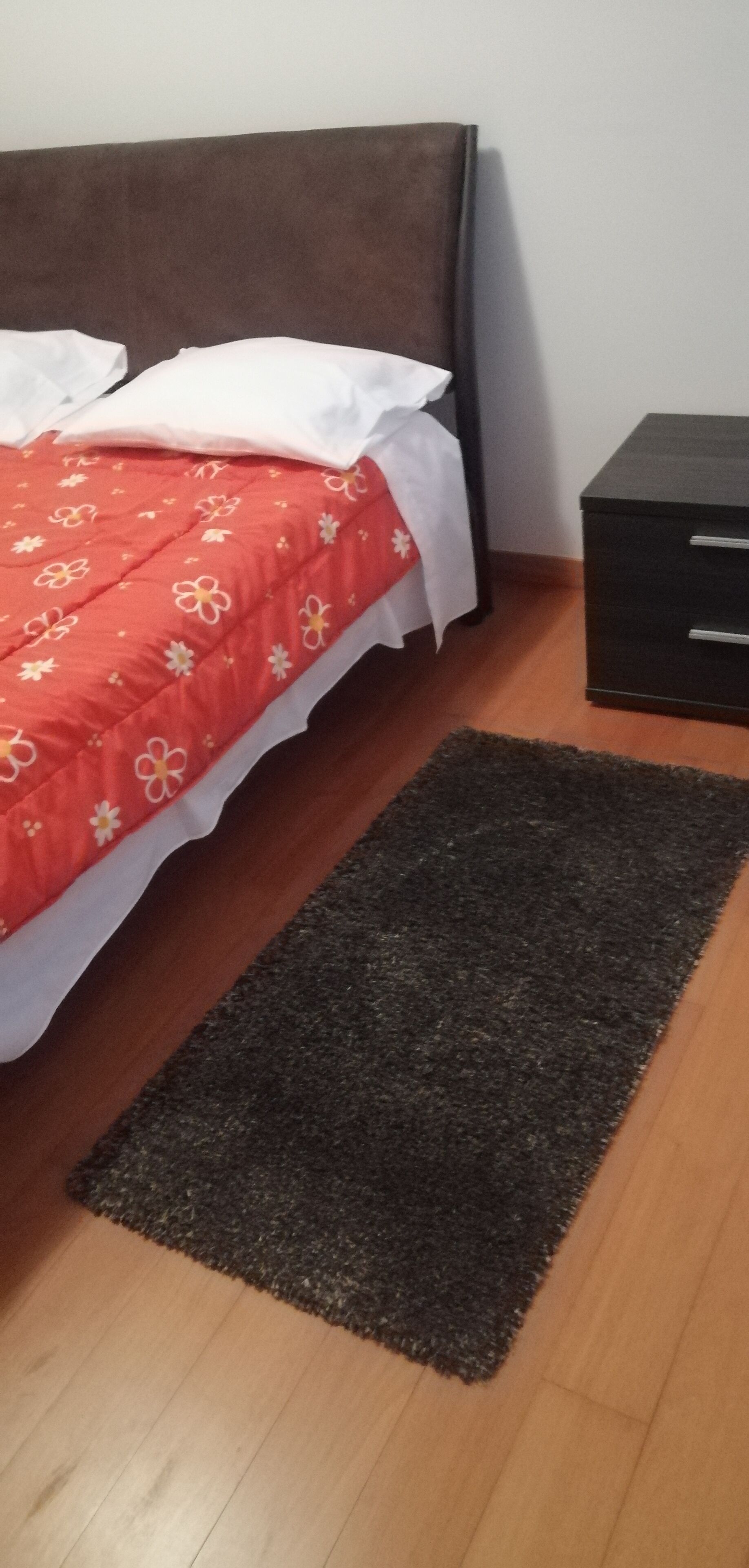 A bed with a red floral bedspread and white pillows, a dark headboard, a black nightstand with drawers, and a dark rug on a wooden floor.